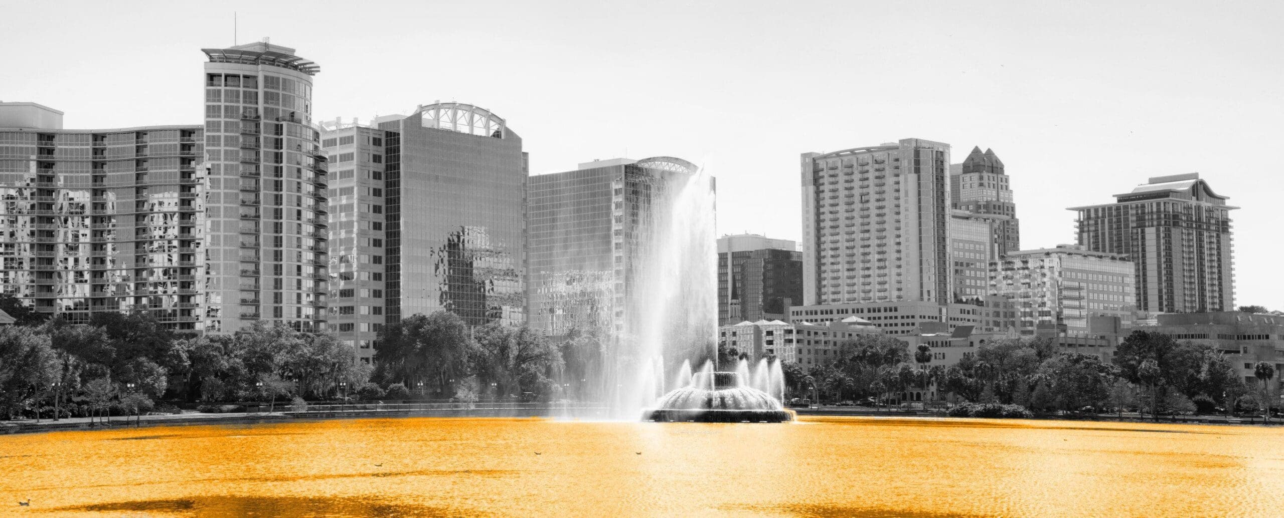 A black and white image of buildings, with a fountain and a lake that's orange