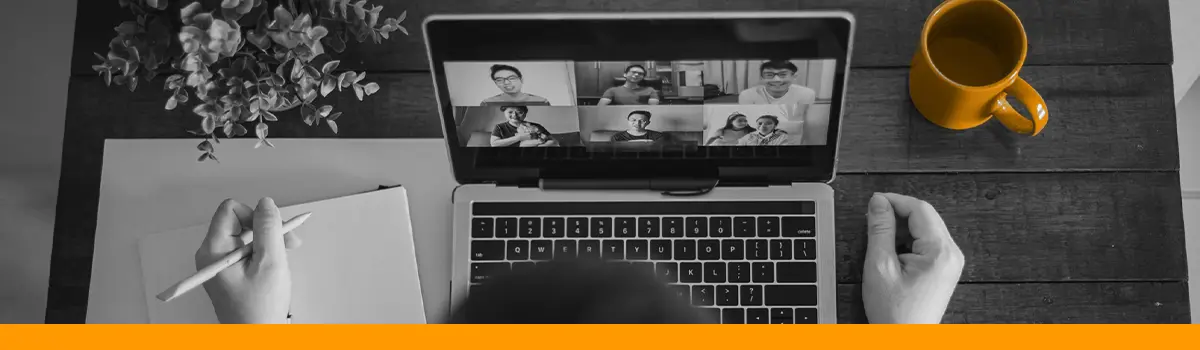 The image shows a person sitting at a desk, viewed from behind, participating in a virtual meeting on a laptop. The laptop screen displays a video conference with six participants in different windows. On the desk are a notebook with a pen in hand, a small potted plant, and a yellow mug, adding a pop of color to the predominantly black-and-white image. An orange bar runs along the bottom edge of the image.